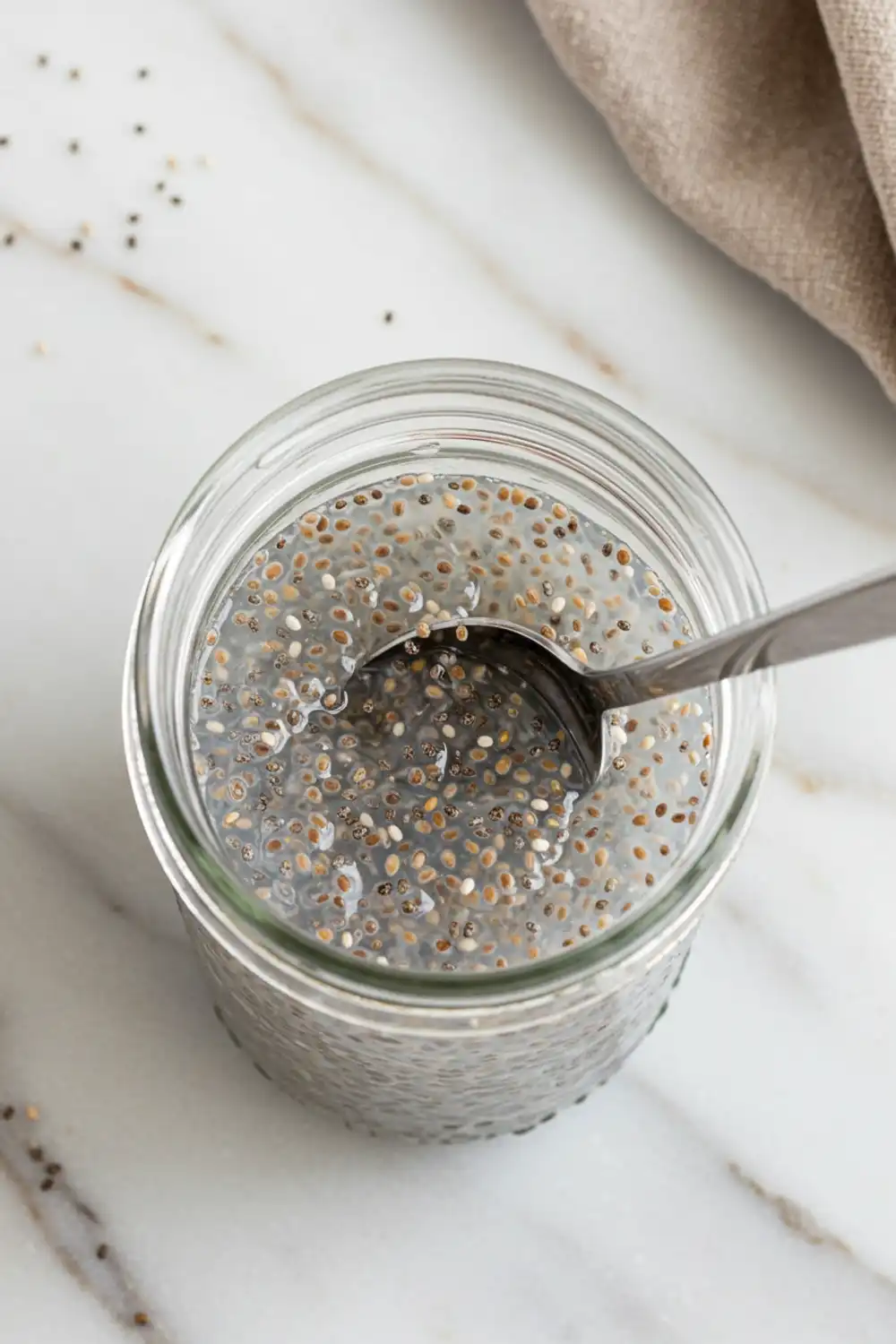 Close-up top-down view of Chia Seeds and Coconut Water Electrolyte Drink assembling in a clear glass jar with subtle wear, showing a spoon stirring chia seeds into coconut water to prevent clumps, with a natural, home-cooked feel.