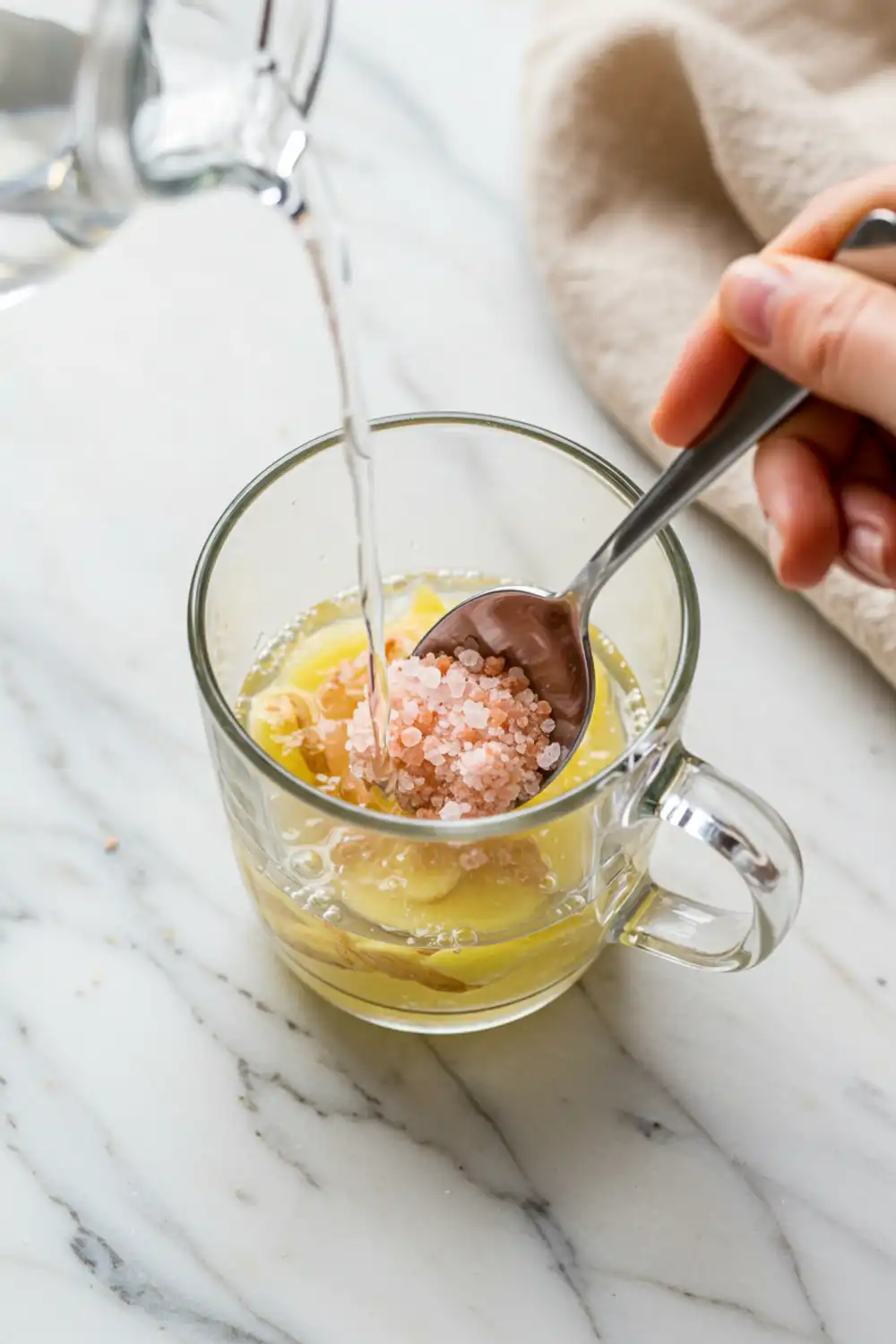 Close-up top-down view of Himalayan Pink Salt and Ginger Detox Drink assembling in a clear mug with subtle wear, showing ginger infusing in warm water and salt dissolving, with a natural, home-cooked feel.