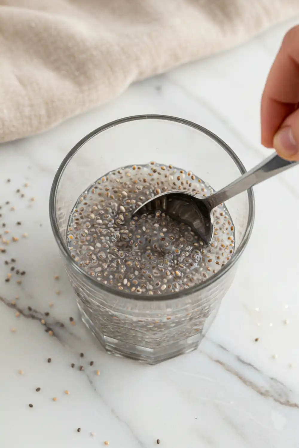 Close-up top-down view of Chia Seed Water assembling in a clear glass with subtle wear, showing chia seeds being stirred in warm water, with a natural, home-cooked feel.