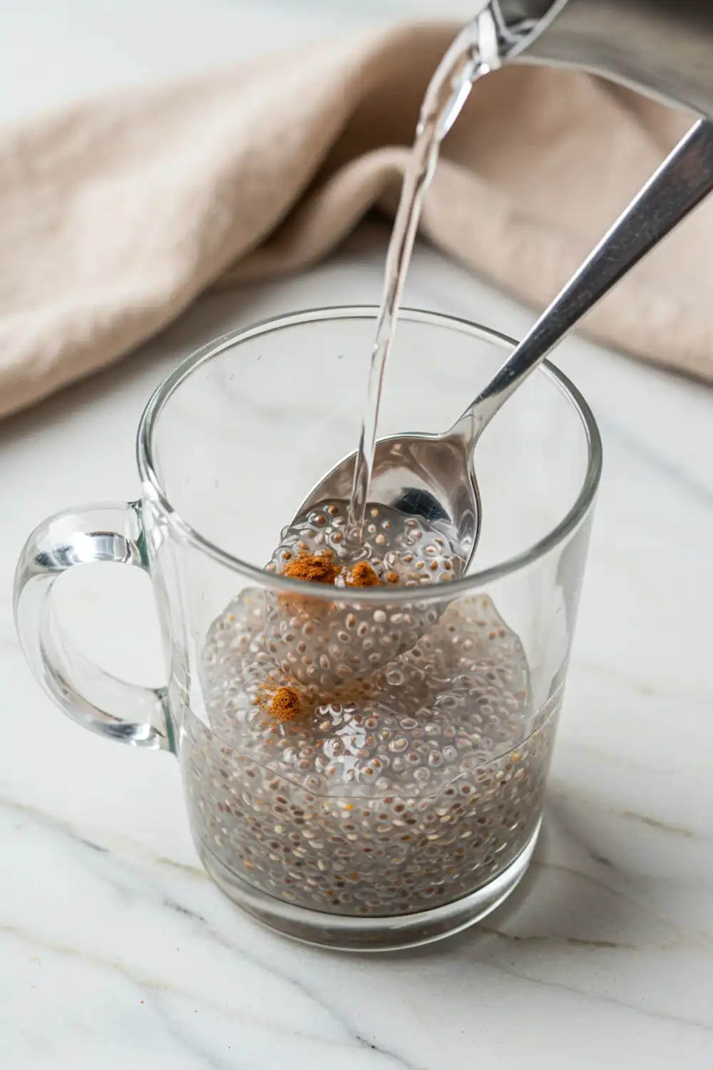 Close-up top-down view of Chia Seed and Cinnamon Water assembling in a clear glass mug with subtle wear, showing chia seeds and cinnamon being stirred with warm water, with a natural, home-cooked feel.
