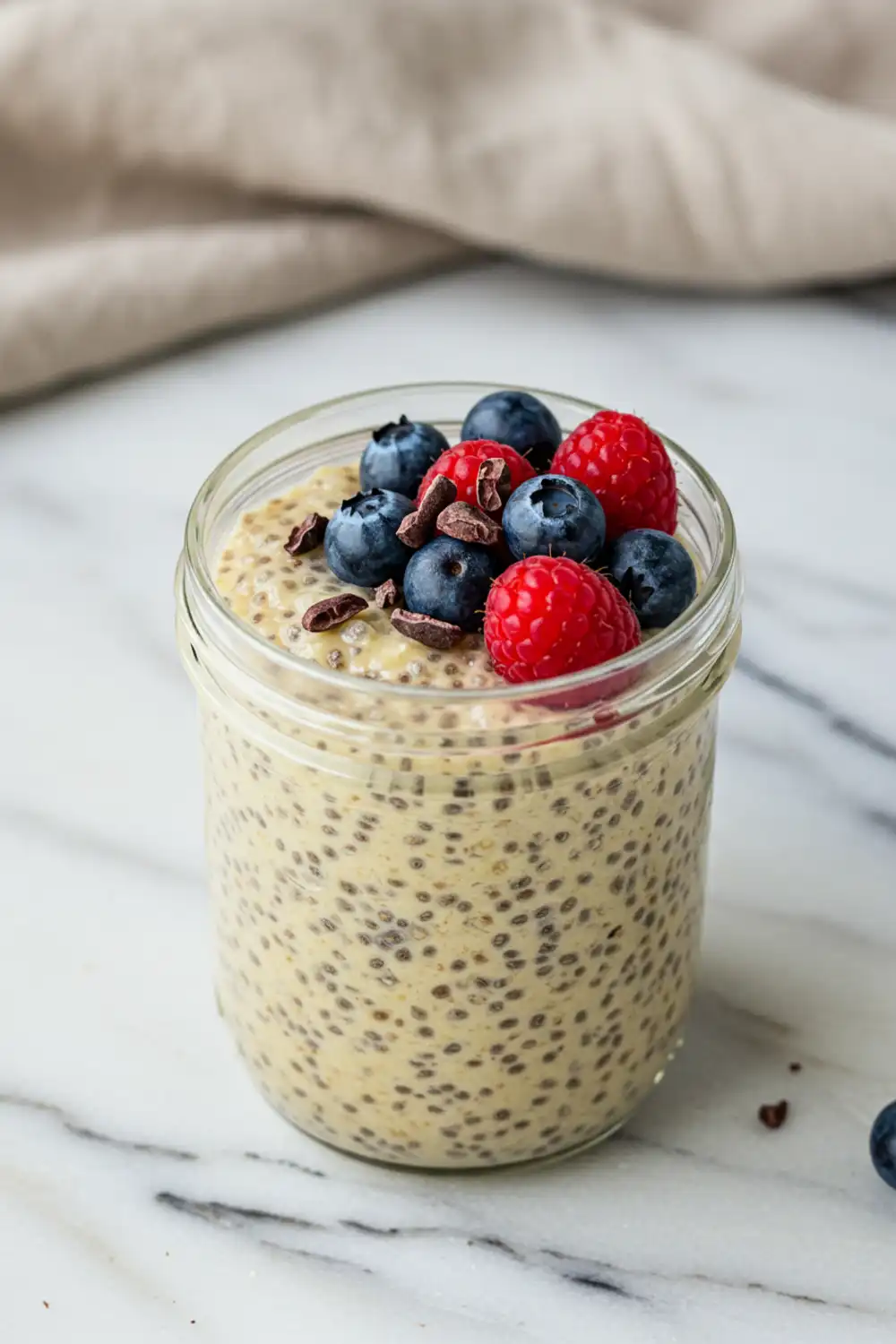 Close-up top-down view of the finished Banana Chia Pudding in a clear glass jar with subtle wear, garnished with fresh mixed berries and cacao nibs, looking natural and home-prepared.